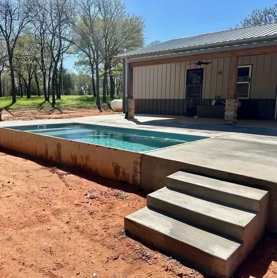Concrete pool with steps next to a covered building, set on dirt.