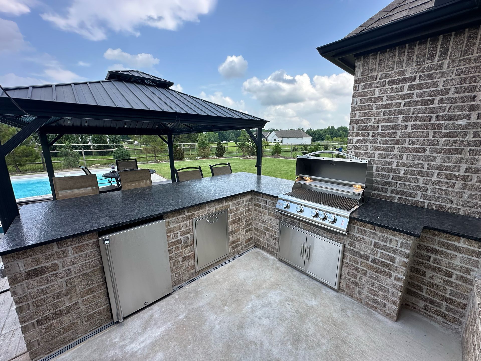 Outdoor kitchen with brick facade, black countertop, grill, and gazebo overlooking a pool on a sunny day.