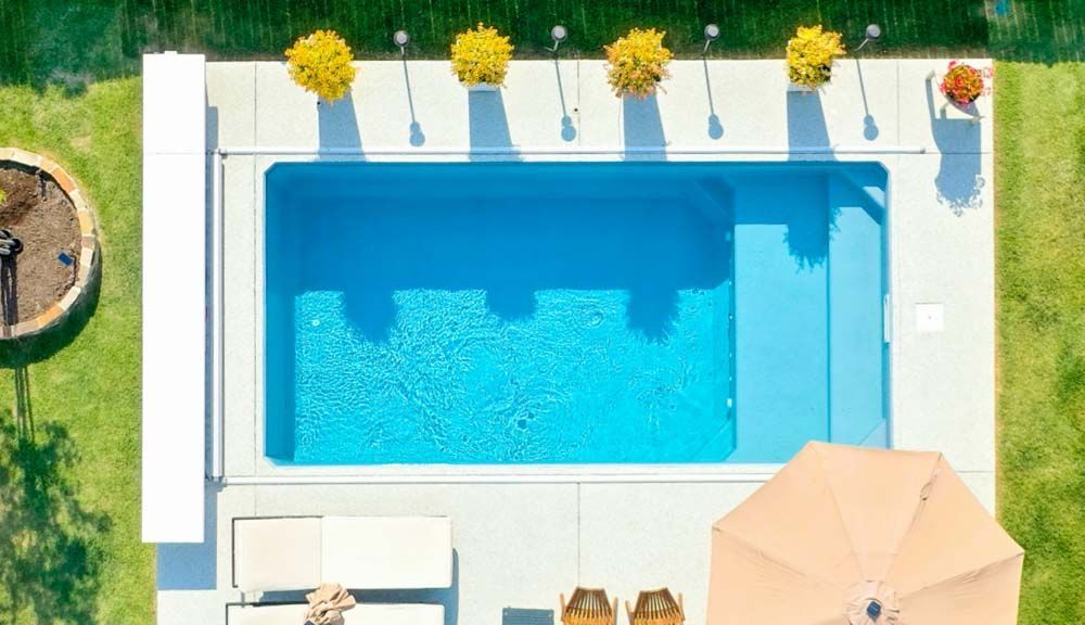 Aerial view of a rectangular blue swimming pool with concrete surround, and a beige patio with seating and umbrella.