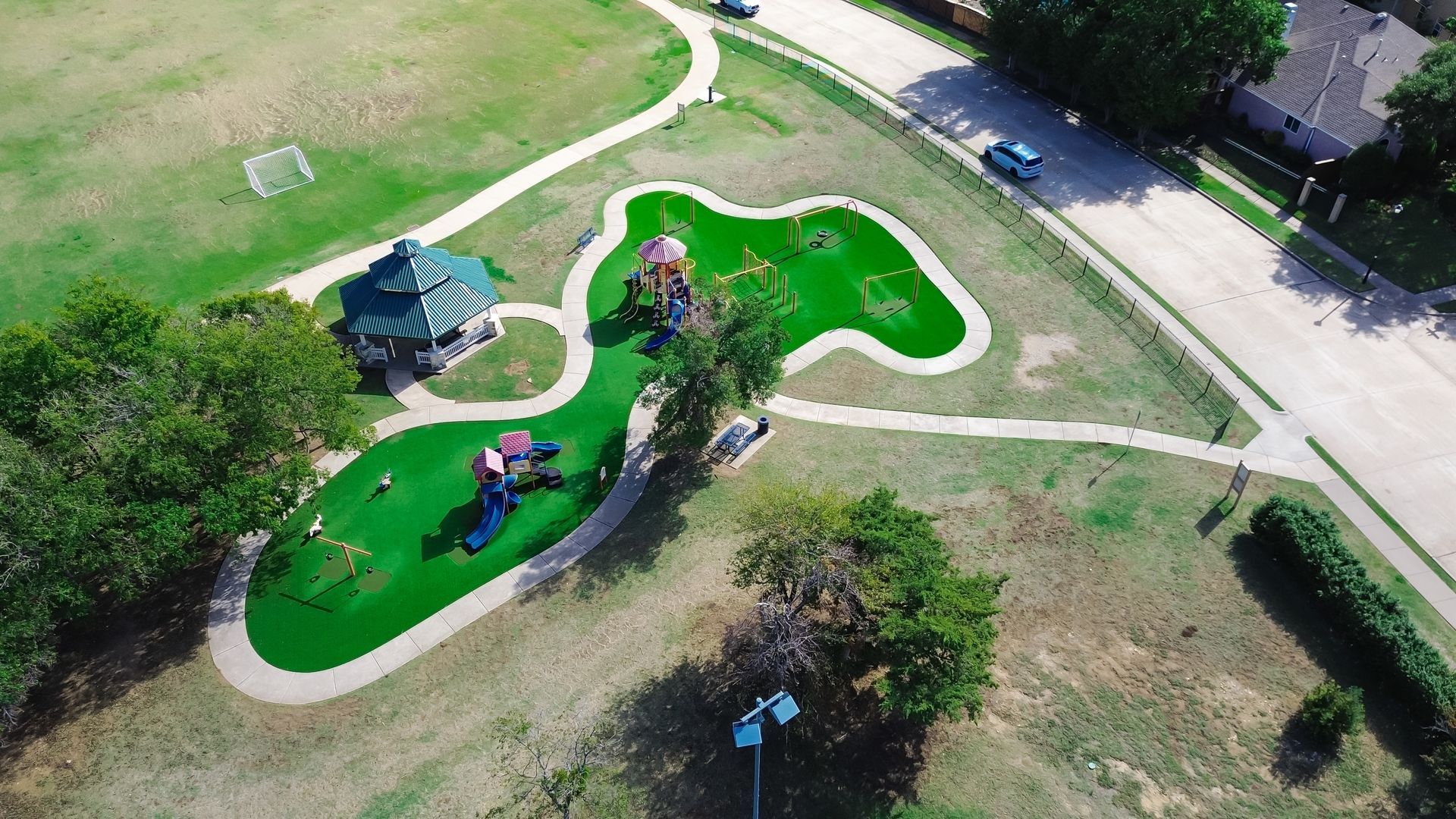 Aerial view of a park with playground equipment on green turf, winding pathways, a gazebo, and a small soccer field.