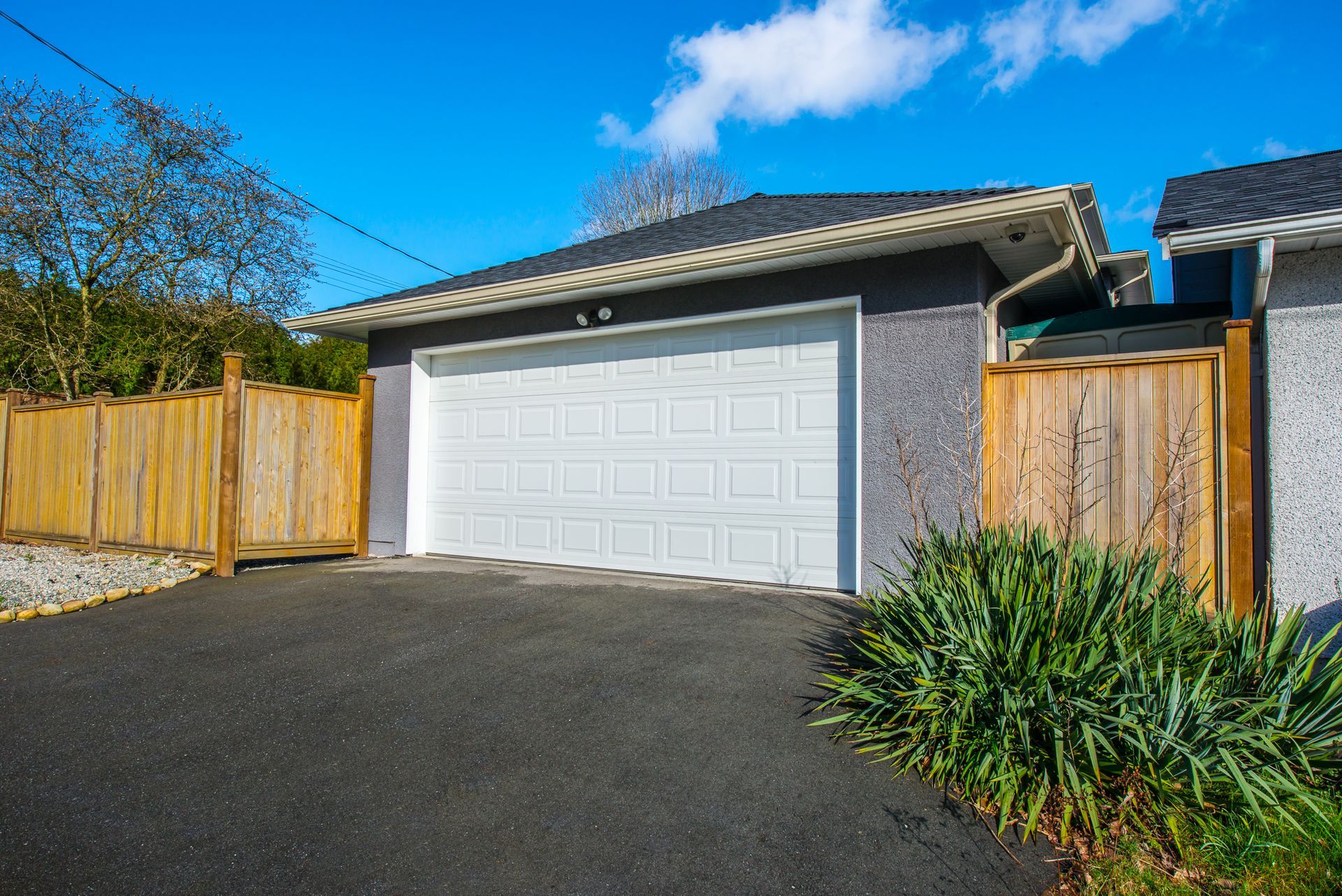 White garage door with black asphalt driveway; wooden fences and green bushes frame it under blue sky.