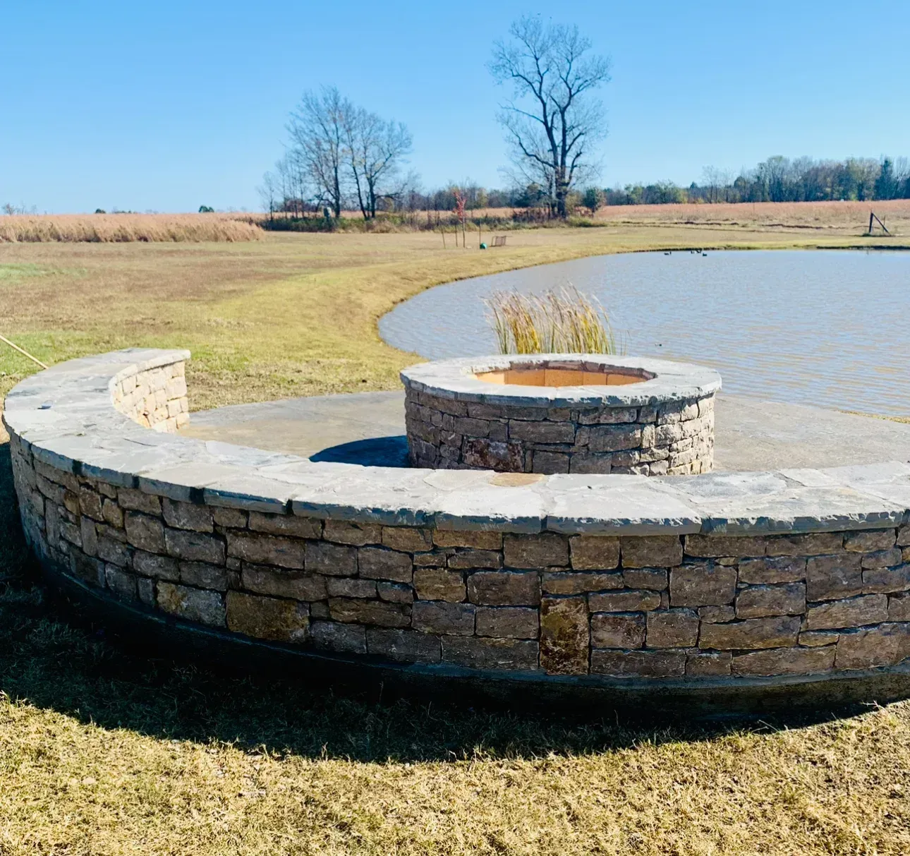 Stone fire pit with surrounding low wall near a pond, fields and trees under a blue sky.