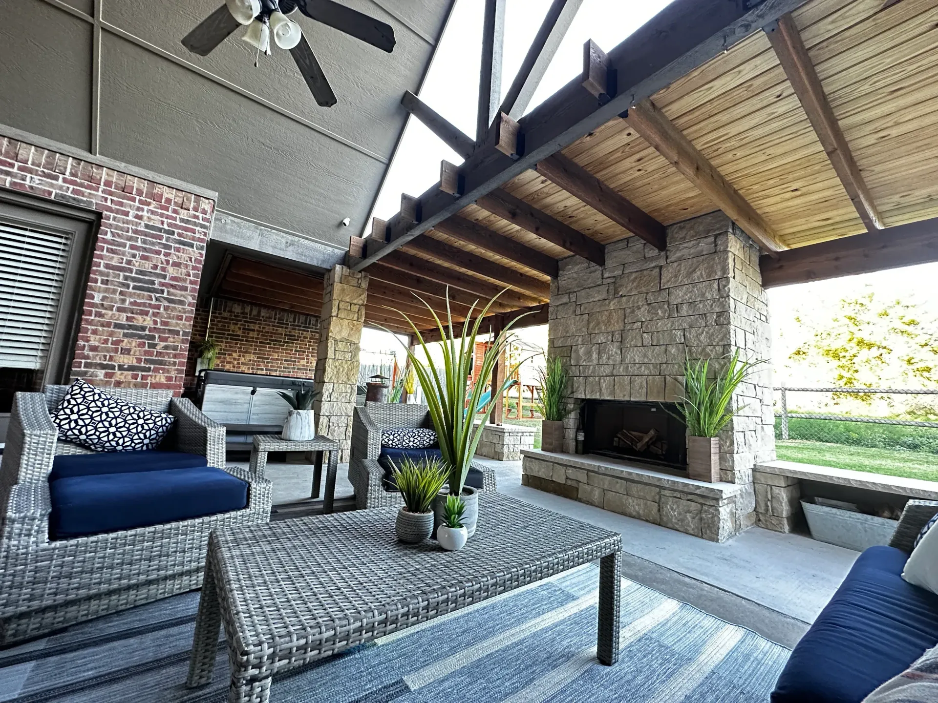 Outdoor living space with a fireplace, seating, brick and stone accents, under a sloped wood ceiling.