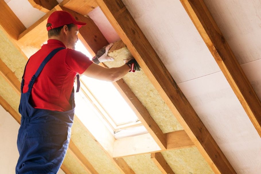 Worker in red shirt and blue overalls installing insulation in a sloped attic ceiling