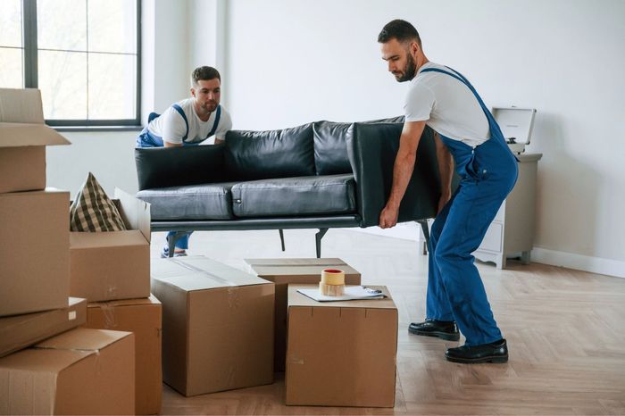 Two movers in blue overalls carry a black couch inside a room with boxes.