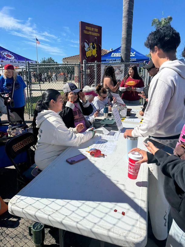 People standing around a long folding table outdoors at an event, with one person holding a red drink cup.