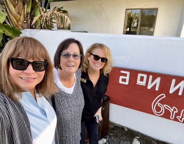 Three smiling individuals stand outside next to a white wall with a red sign reading