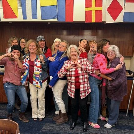 A group of eleven people standing in a row against a wall decorated with nautical flags, smiling and making heart signs.