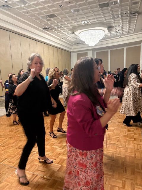 A group of people dancing on a wooden floor in a ballroom with a large chandelier overhead.
