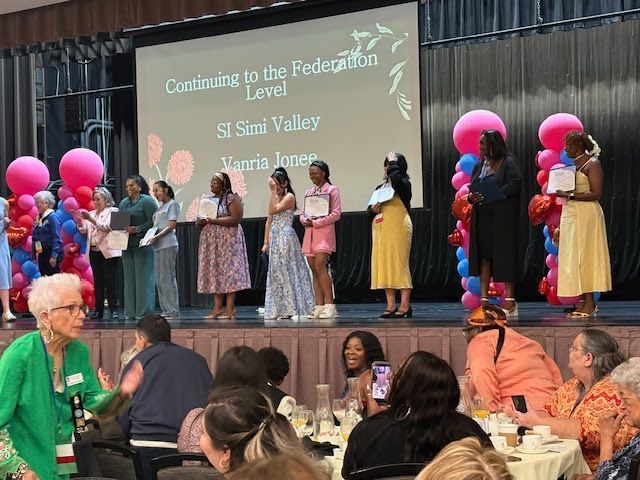A group stands on a stage before a screen announcing winners moving to the Federation level, with audience members below.