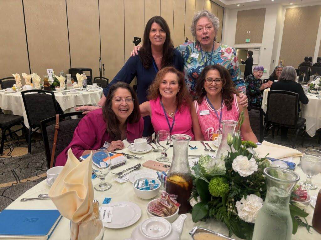 Six smiling people pose for a group photo around a dining table at a conference or banquet event.