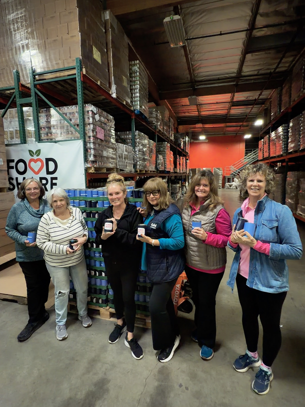 Six volunteers in a Food Share warehouse stand in a line, each holding a small blue product, smiling toward the camera.