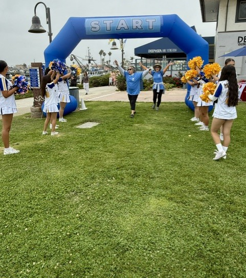 Blue START archway at a grassy outdoor event with cheerleaders and spectators lined along the sides. Women's Walk and Expo on Saturday, May 9, 2026.
