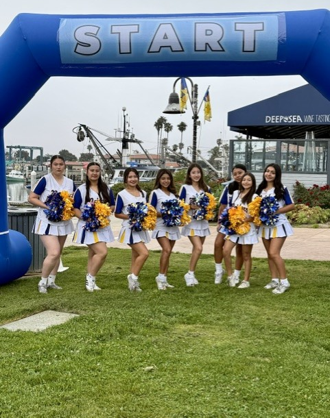 Cheer squad posing under blue START arch at a harbor with boats and a DHS sign in the background. Women's Walk and Expo on Saturday, May 9, 2026.