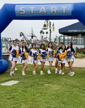 Cheer squad posing under blue START arch at a harbor with boats and a DHS sign in the background. Women's Walk and Expo on Saturday, May 9, 2026.