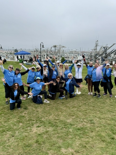People in blue shirts and masks pose with arms raised on a grassy field near the water. Women's Walk and Expo on Saturday, May 9, 2026.