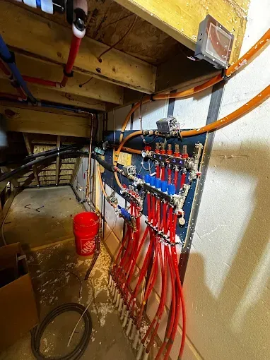 Underneath a wood structure, red and blue radiant floor heating pipes connect to a manifold.