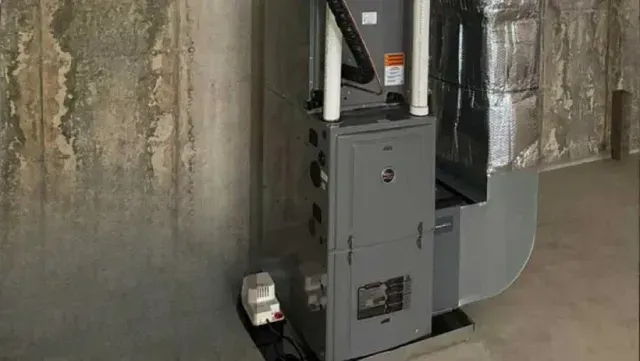 Furnace in a basement; gray metal unit, silver ductwork, next to a concrete wall.
