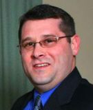 Man in glasses smiling, wearing a blue shirt, tie, and dark suit jacket.