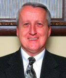 Man in a dark suit smiles, wearing a tie; light complexion, short gray hair.