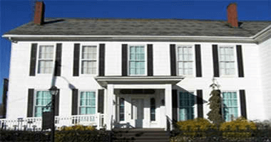 White two-story house with black shutters, front porch, gray roof, and brick chimneys against a blue sky.