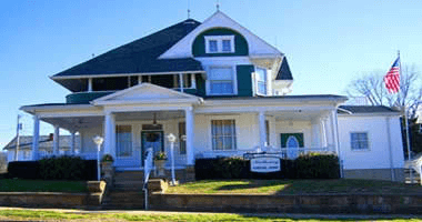 White Victorian house with green trim, porch, and American flag on a sunny day.