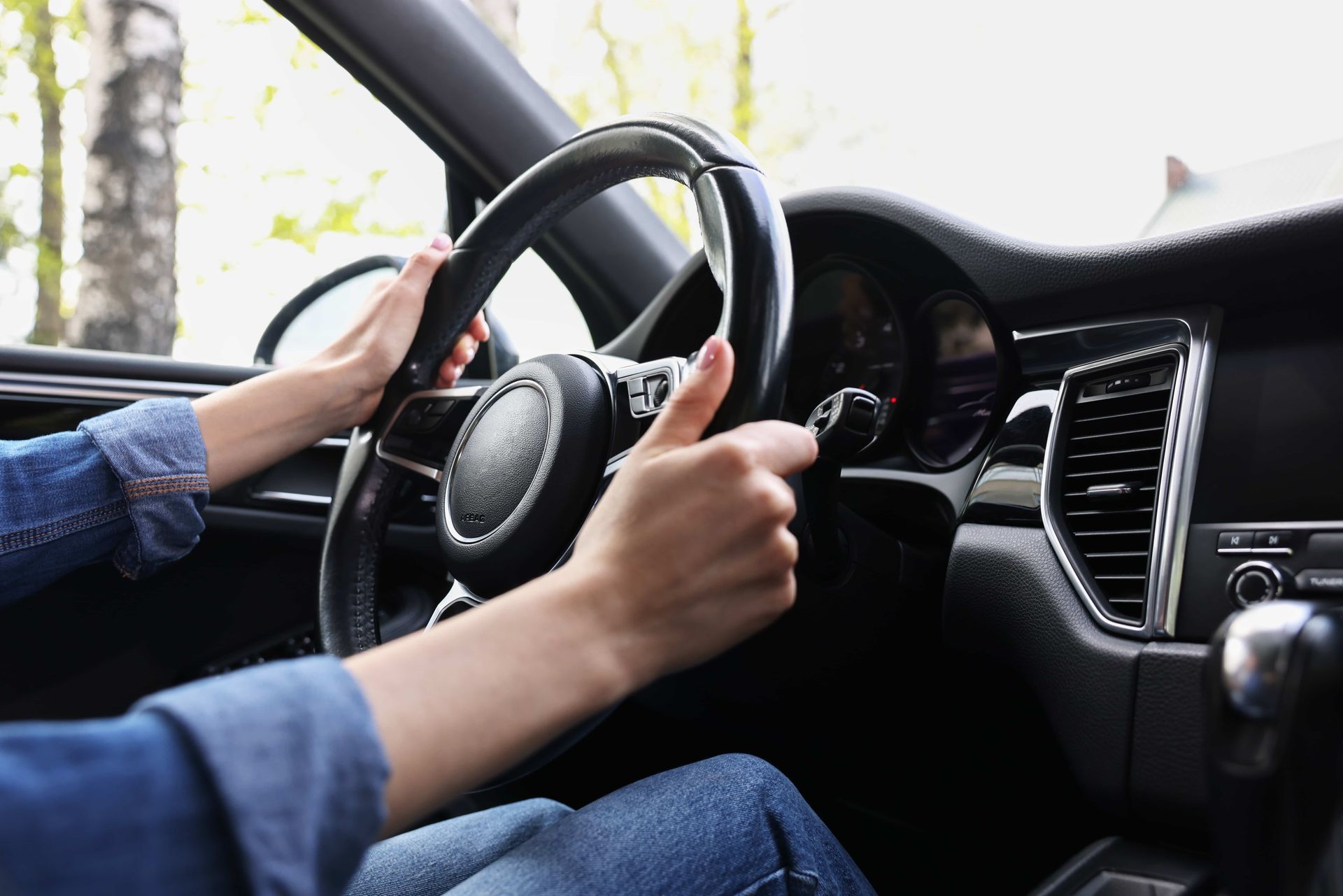 Person's hands gripping a car's steering wheel, driving. Denim sleeves and a dark dashboard are visible.