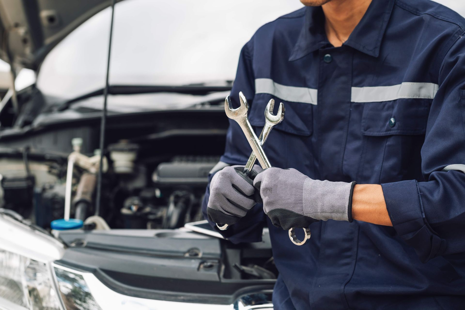 Mechanic holding wrenches near a car engine, wearing a blue jumpsuit and gloves.