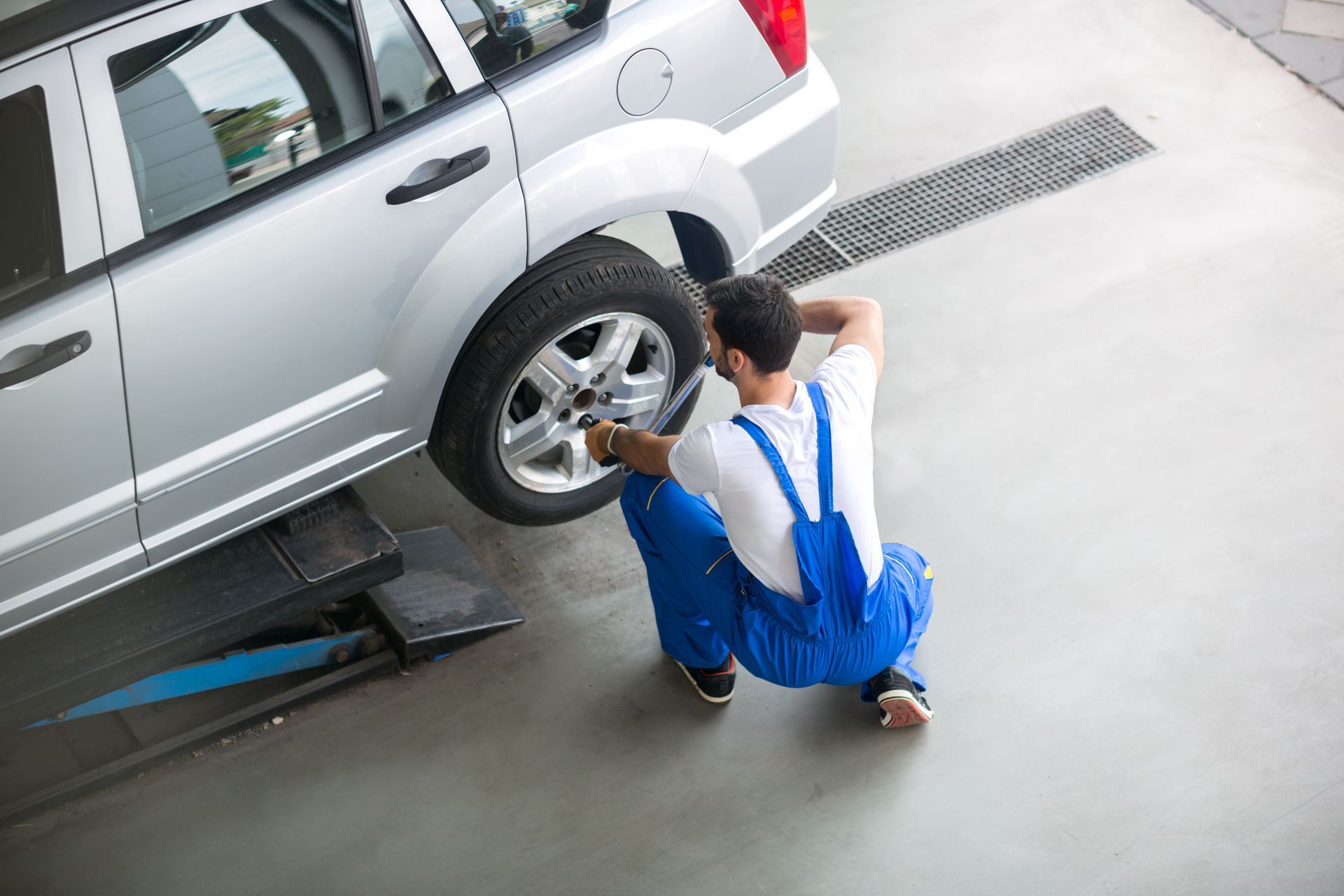 Mechanic in blue overalls removing a tire from a silver car in a garage.