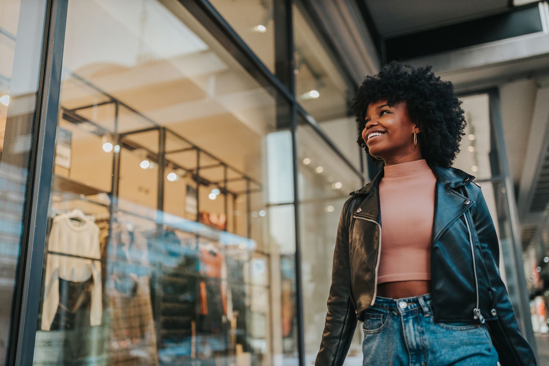 Jeune femme souriante portant une veste en cuir, marchant devant une vitrine de magasin.