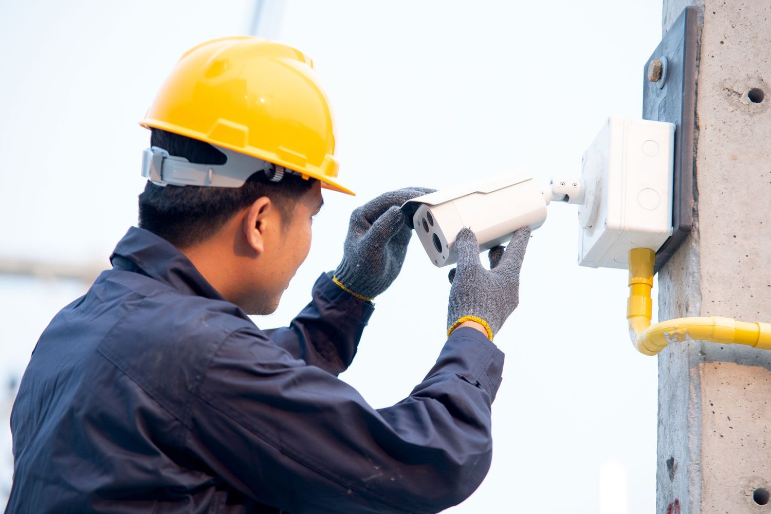 A worker in a blue uniform and yellow helmet, performing CCTV installation in an outdoor setting.