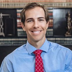 Man in blue shirt and red tie smiling, with bookshelves in the background.