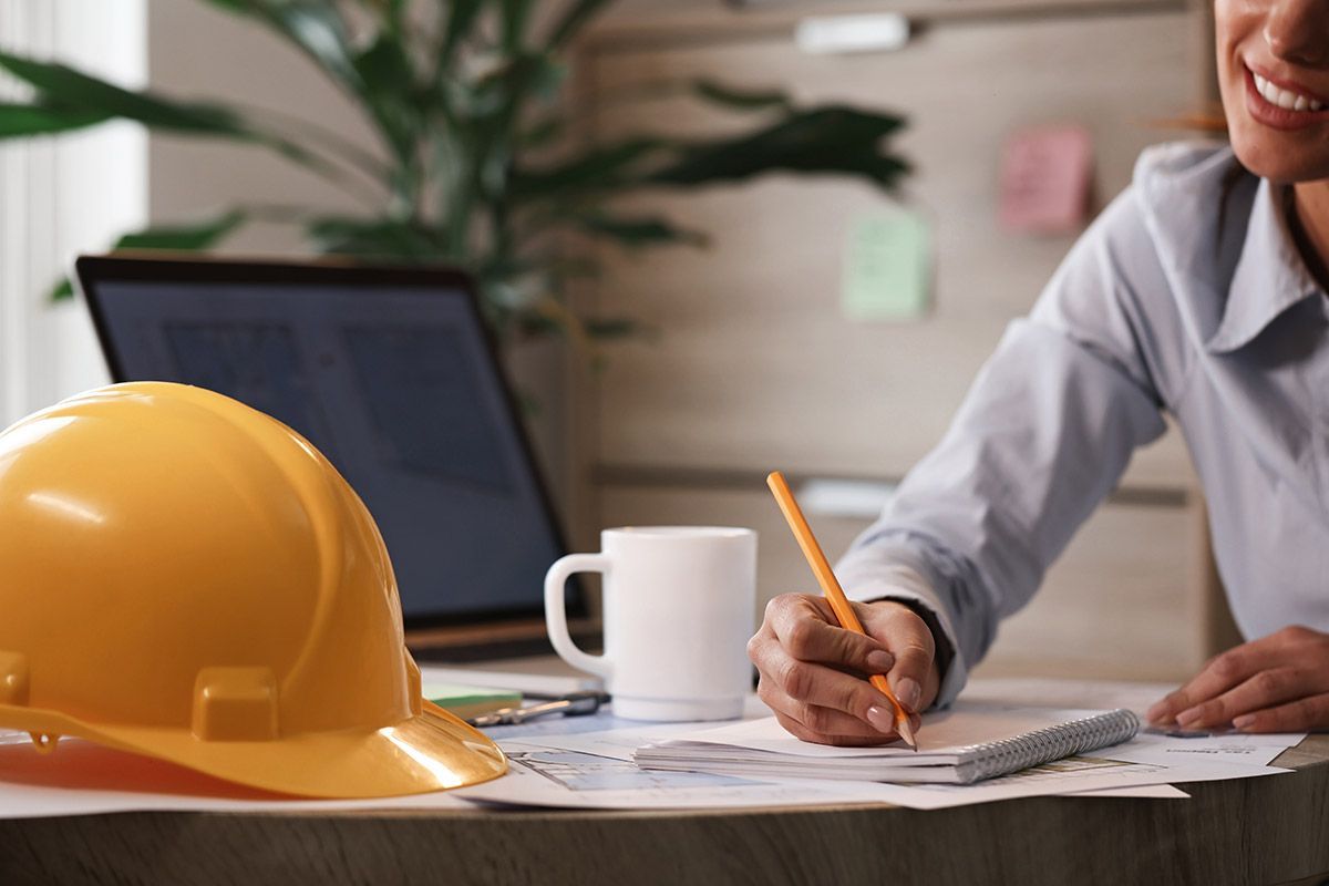 Engineer writing notes beside yellow hard hat and laptop in an office setting