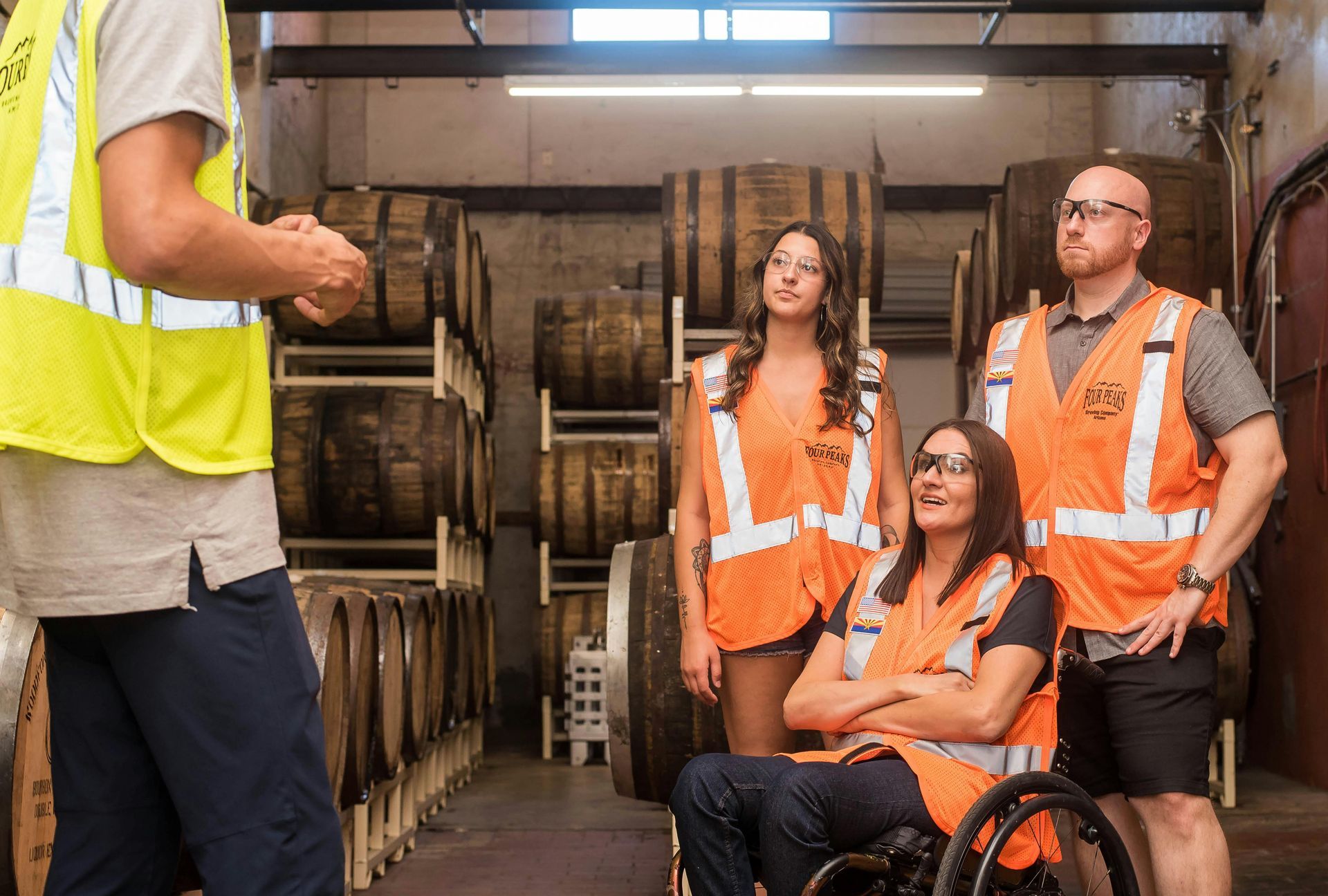 A group of people in safety vests are standing in a warehouse . one of the people is in a wheelchair.