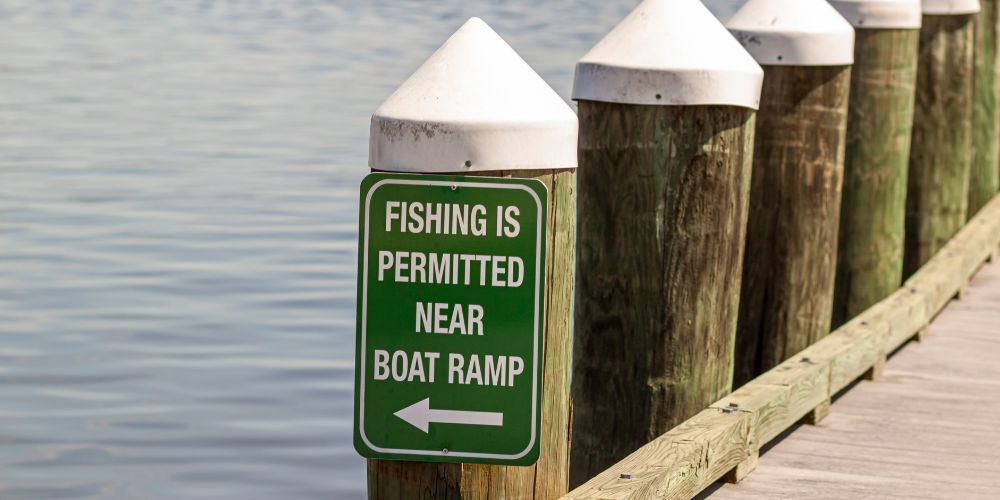 Sign indicating fishing permitted near boat ramp, green sign on wooden post by water.