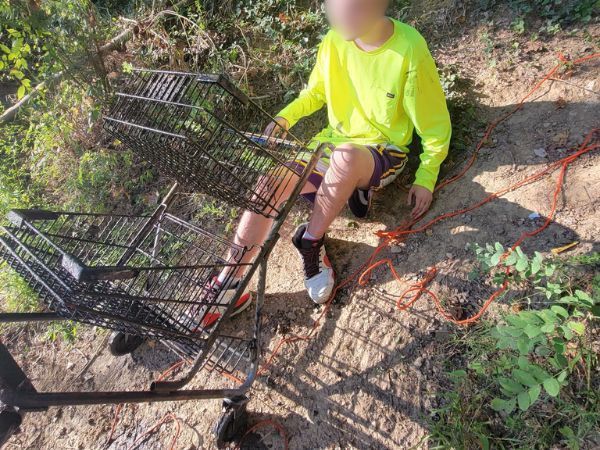 Person sitting next to two overturned shopping carts in a wooded area.  Orange rope present.