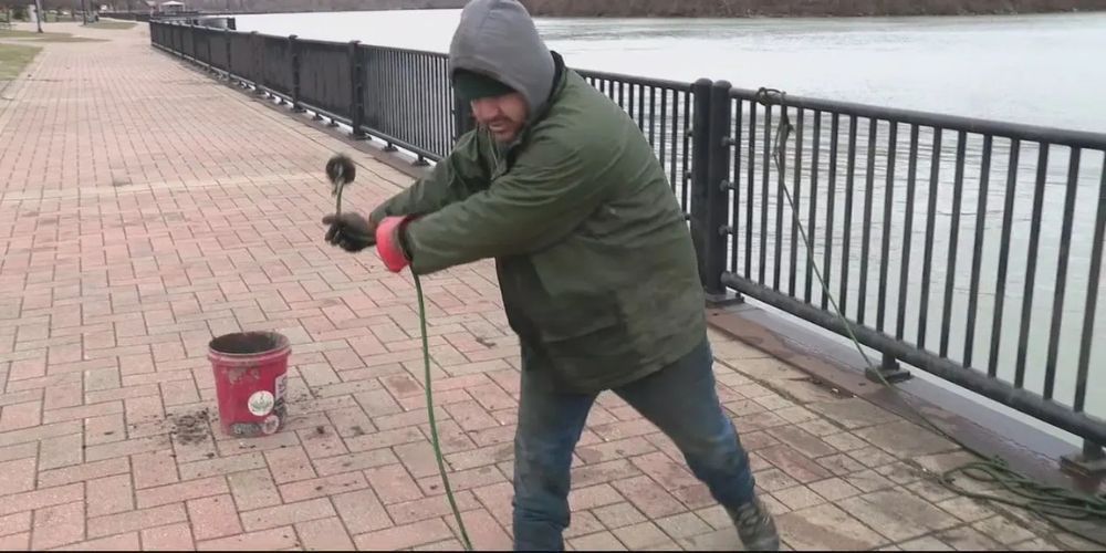 A man is standing on a sidewalk next to a lake swinging a magnet on a rope.