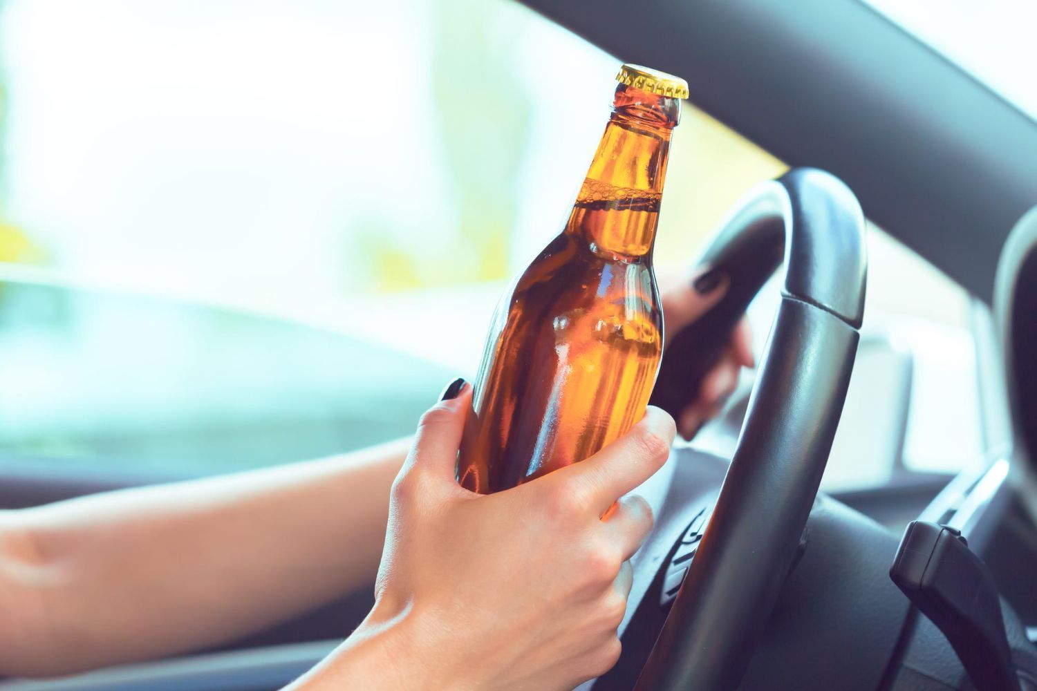 A person’s hand holds a brown glass bottle while gripping a car steering wheel.