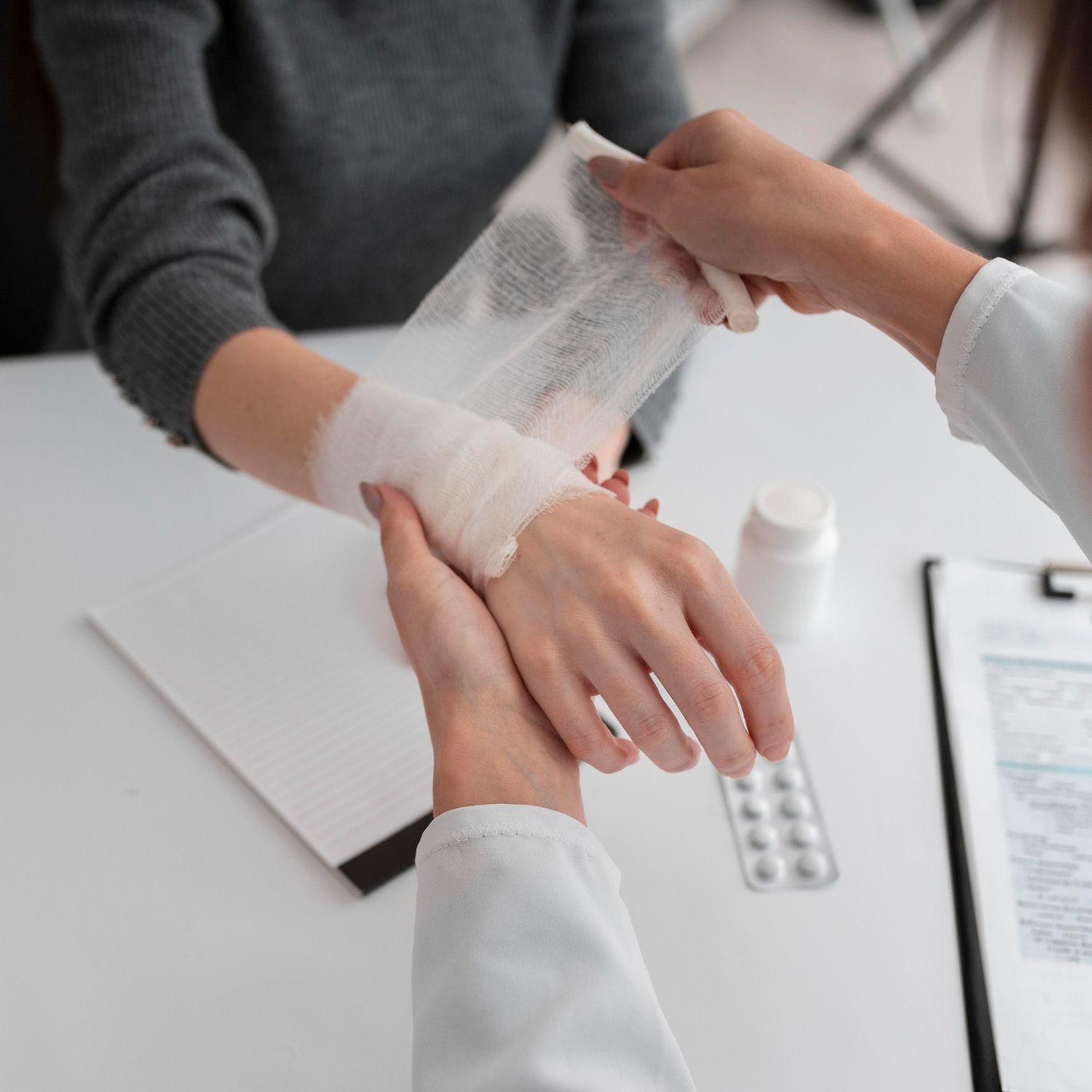 A person in a medical coat applies a gauze bandage to another person's wrist in a sterile office setting.