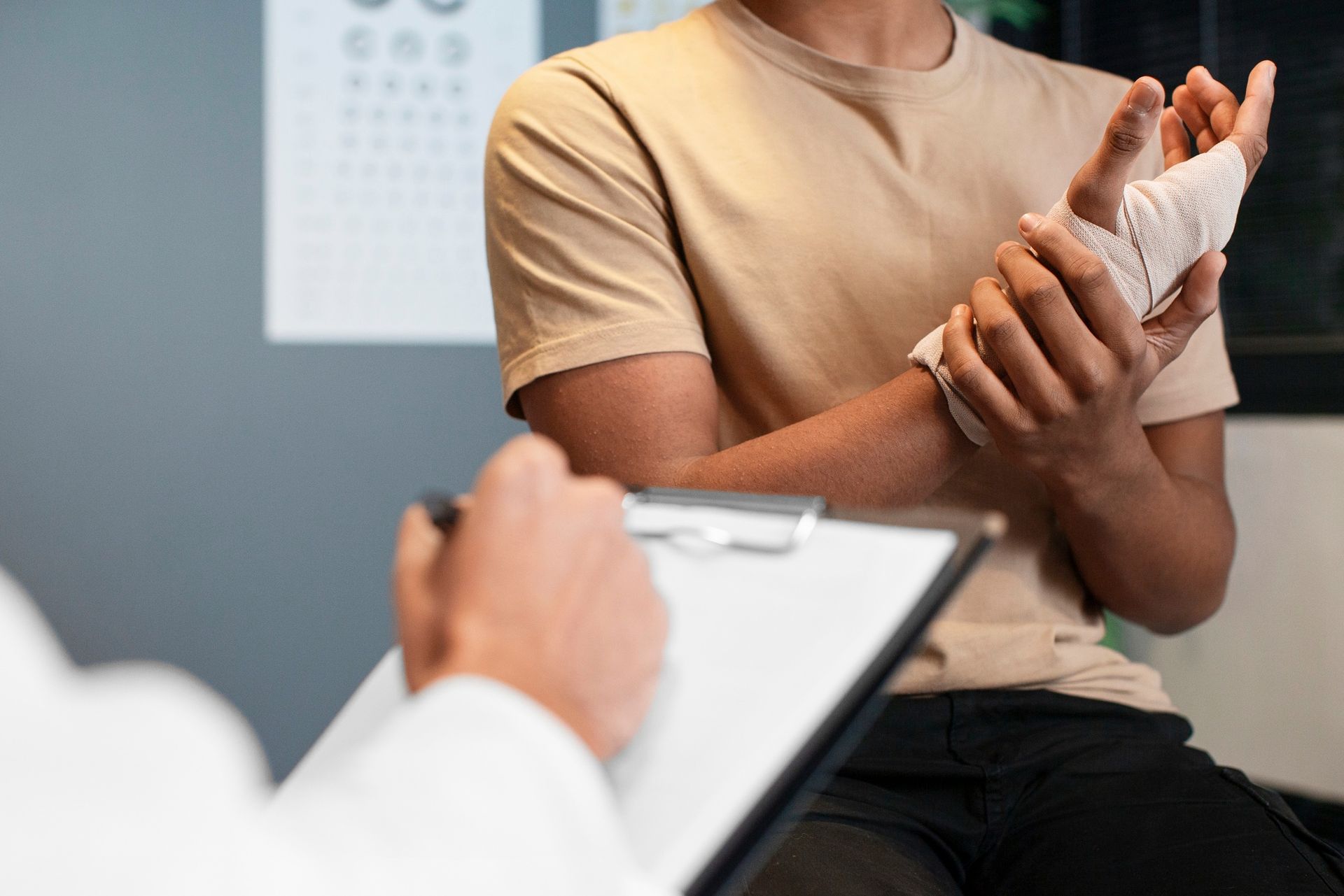 A person with a bandaged wrist sits in a clinic while a medical professional records notes on a clipboard.