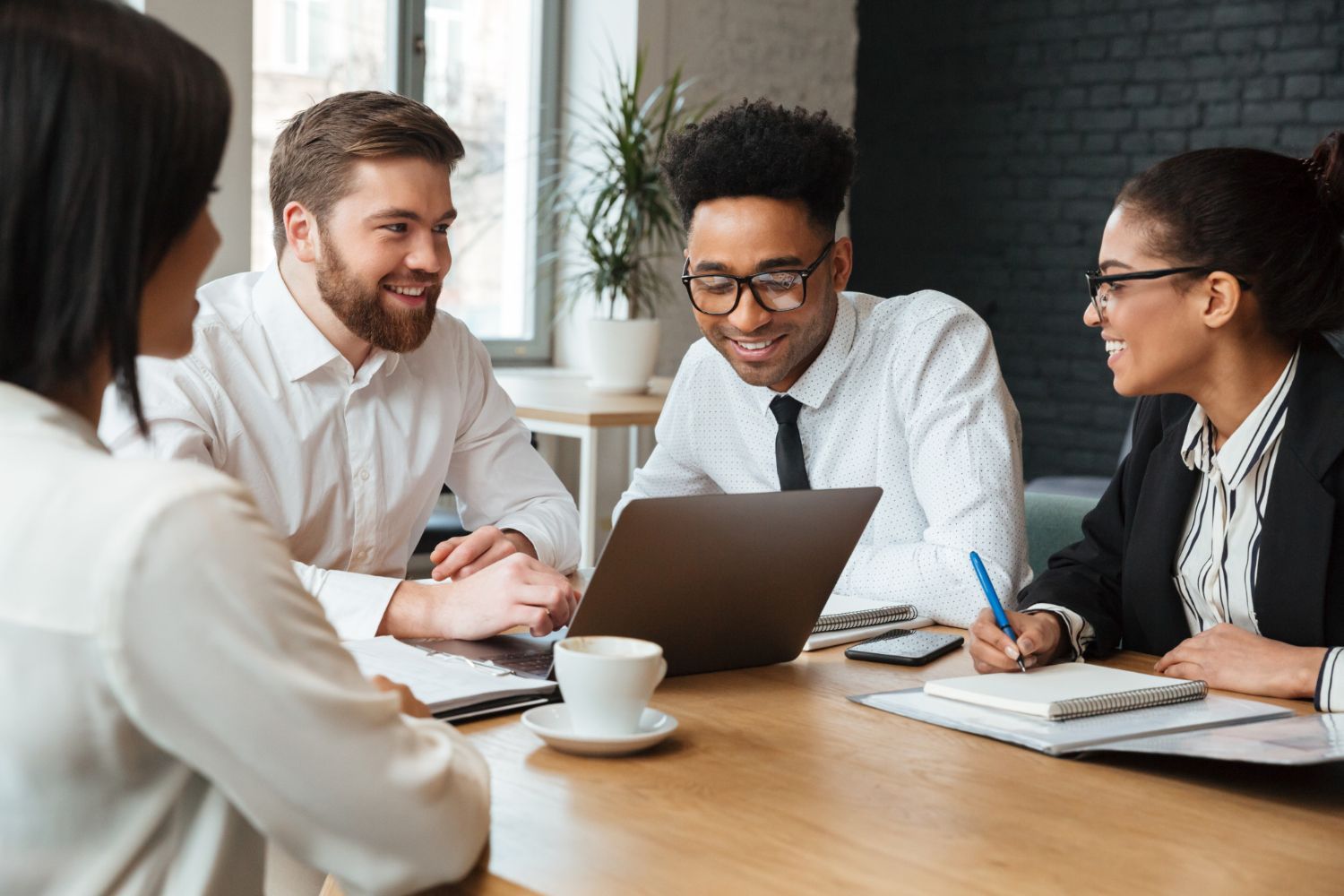 Four professionals in business attire collaborate around a wooden table, focusing on a laptop during an office meeting.