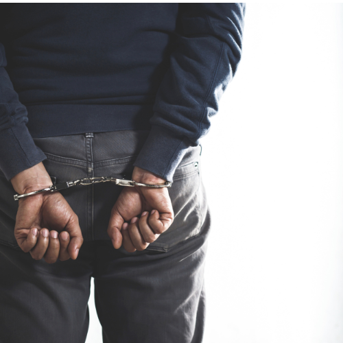 A person from the waist down wears a blue sweater and gray pants, with their hands cuffed behind their back.