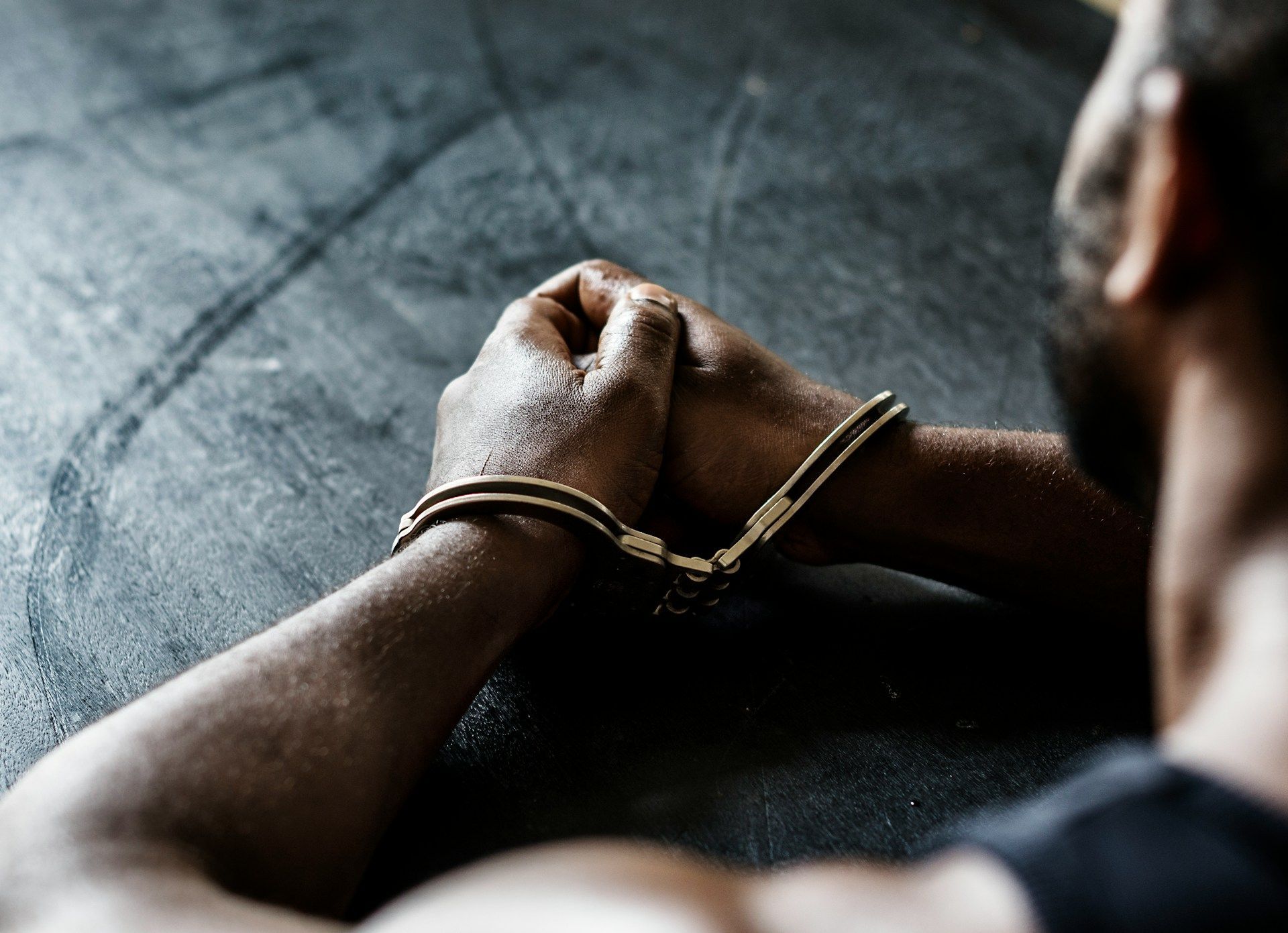 A person sits with their hands restrained by metal handcuffs on a dark, textured surface.