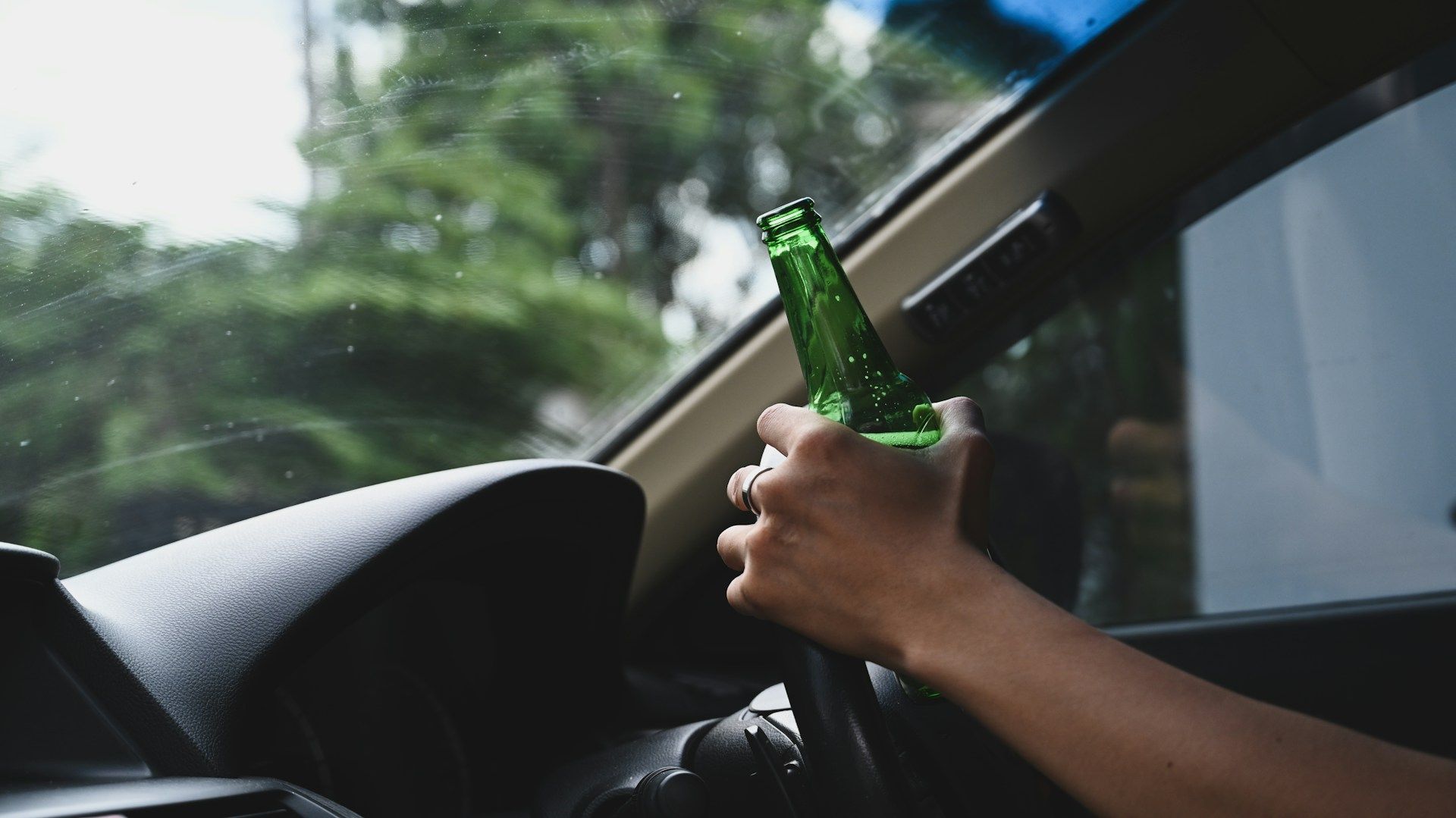 A driver’s hand holds a green glass beer bottle while gripping the steering wheel inside a car.