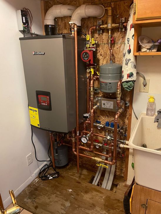 A grey wall-mounted boiler unit with copper piping, a red pump, and a grey expansion tank in a basement utility room.