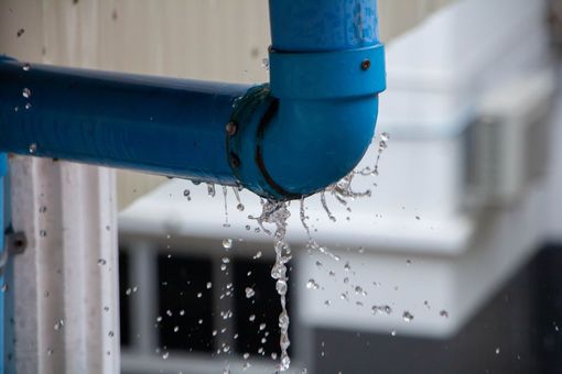 A blue, L-shaped pipe joint leaking water in a steady stream against a blurred background.