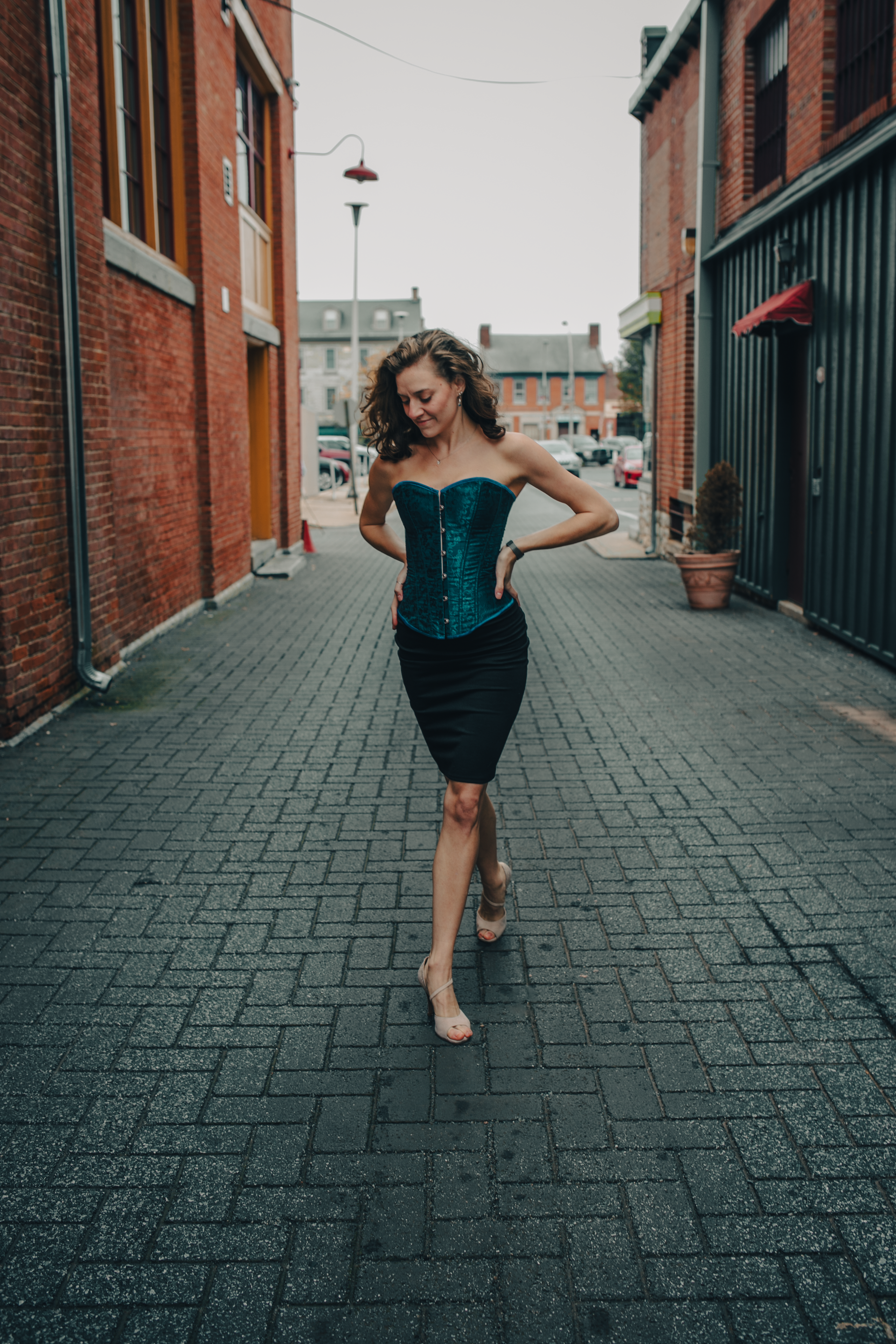 Woman in teal corset and black skirt smiles, poses in brick alleyway.