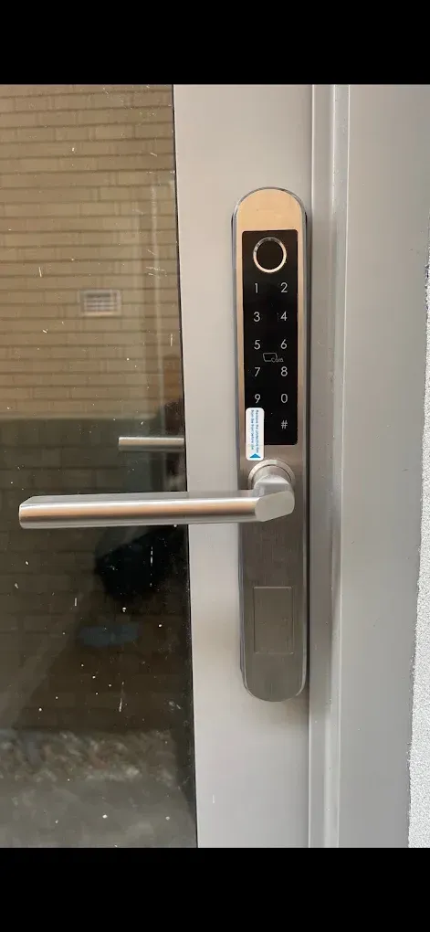 A silver smart lock with a keypad and fingerprint scanner is mounted on a glass door.
