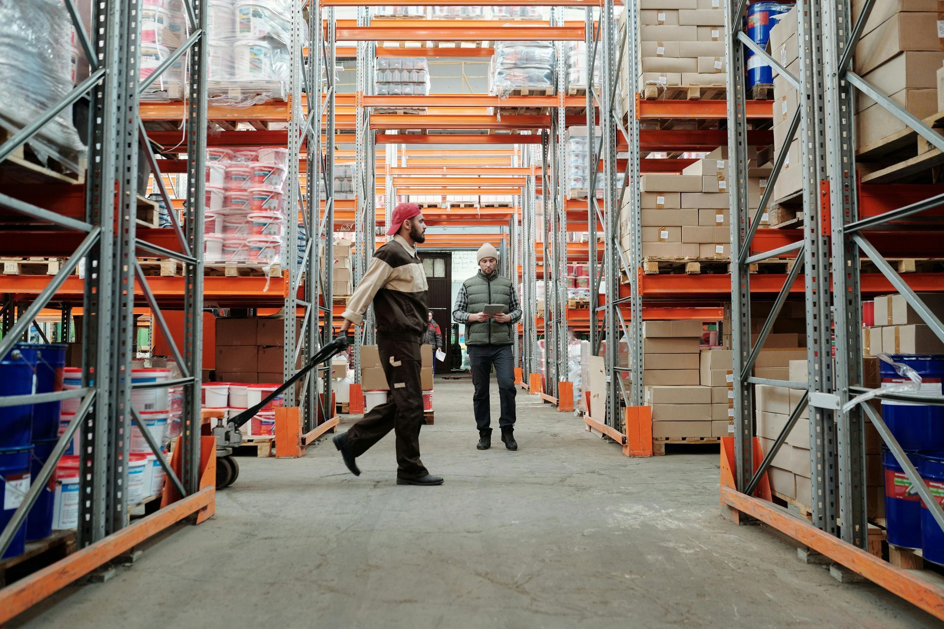 Warehouse scene: person with pallet jack, another walking, shelves of goods.