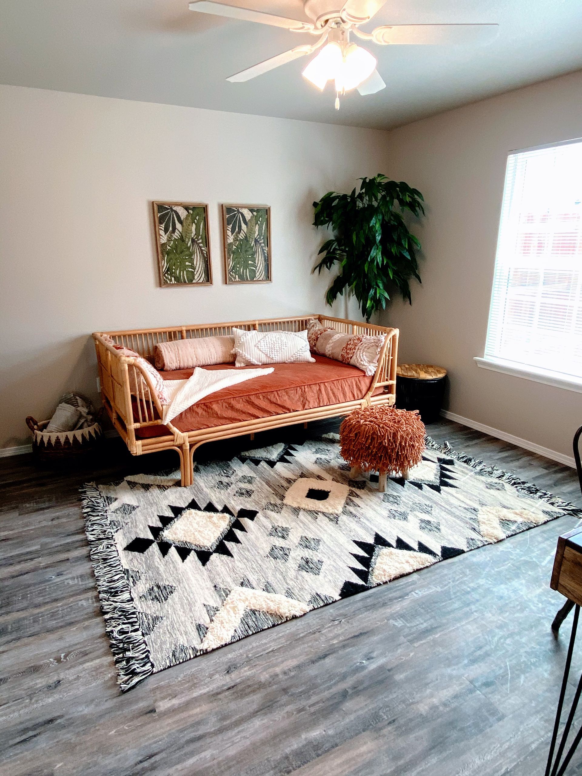 Living room with a boho style: rattan daybed, patterned rug, plants, framed art, and a ceiling fan.